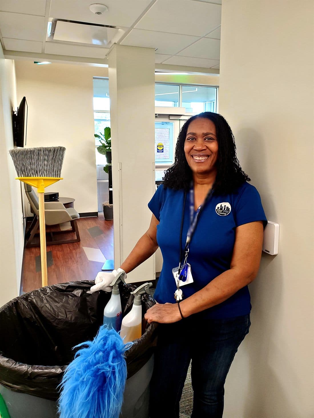 A Your Way Cleaning employee in a blue company polo, smiling beside her cart of supplies in a client's office corridor.